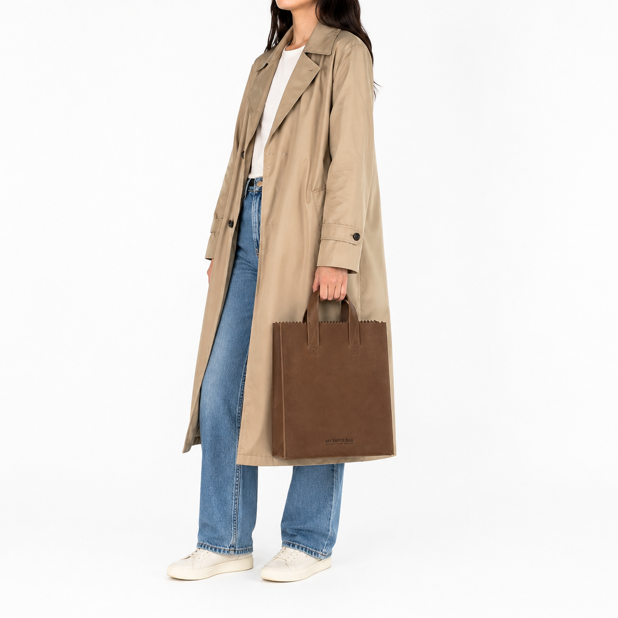 Person wearing a beige trench coat and blue jeans, holding a brown tote bag on a white background