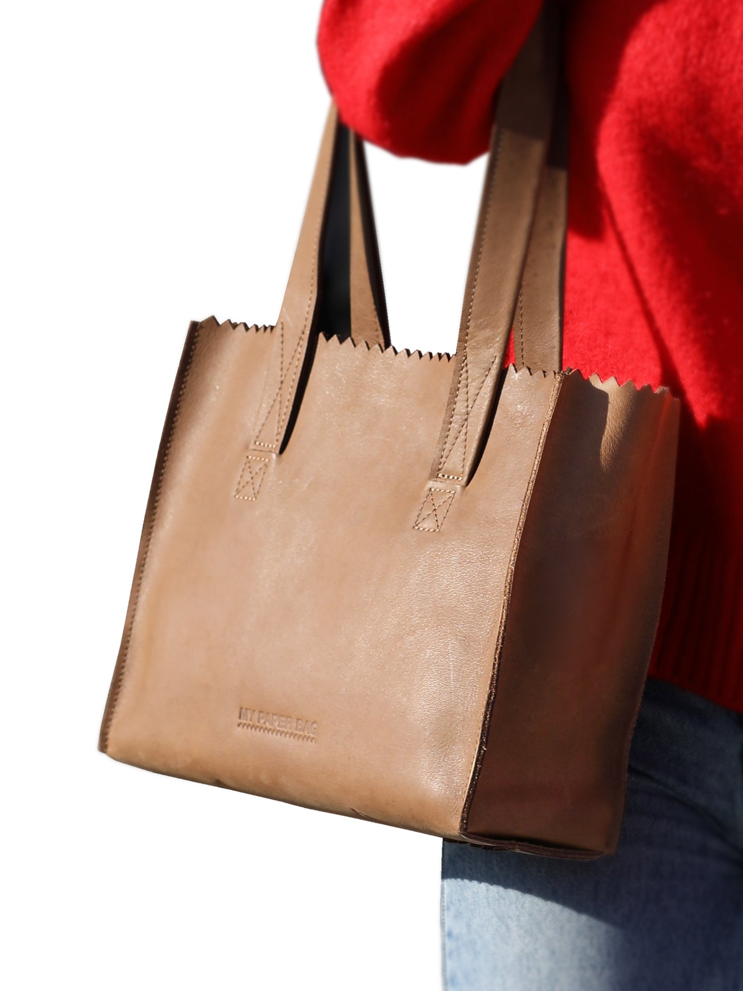 Brown leather tote bag held by a person wearing a red sweater and blue jeans on a white background