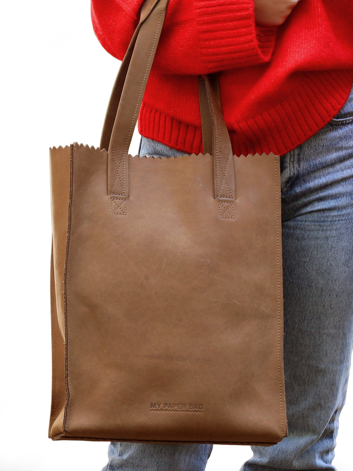 Brown tote bag held by a person wearing a red sweater and blue jeans on a white background
